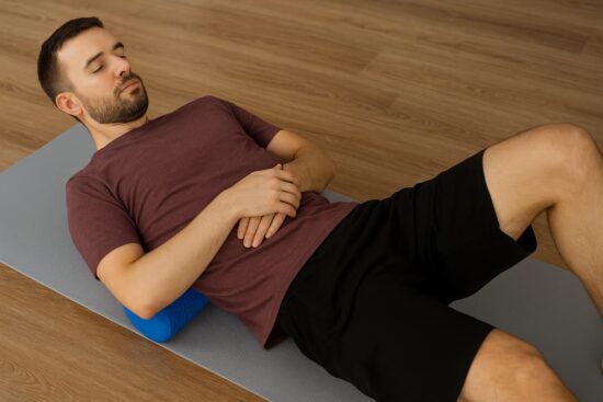 Liberación miofascial con rodillo de espuma para aliviar la tensión y mejorar la flexibilidad Hombre realizando liberación miofascial con un rodillo de espuma azul en la parte superior de la espalda, acostado sobre una colchoneta gris en un suelo de madera.