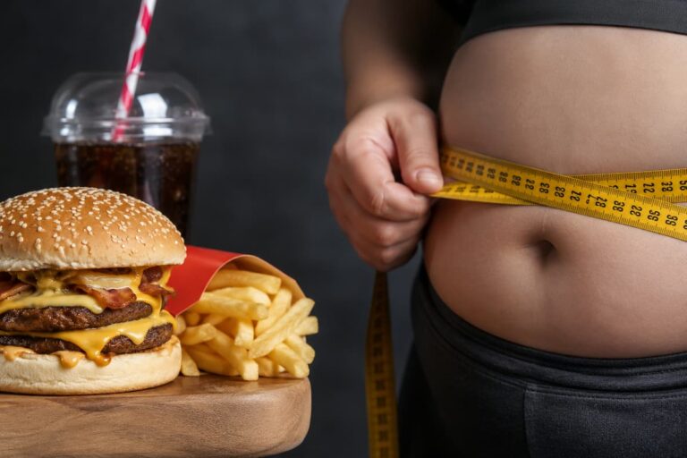 Persona midiendo su cintura con una cinta métrica junto a una hamburguesa, papas fritas y una bebida azucarada.